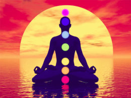 Silhouette of a man meditating with seven colorful chakras upon ocean by red sunset