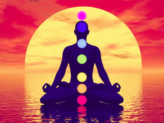 Silhouette of a man meditating with seven colorful chakras upon ocean by red sunset